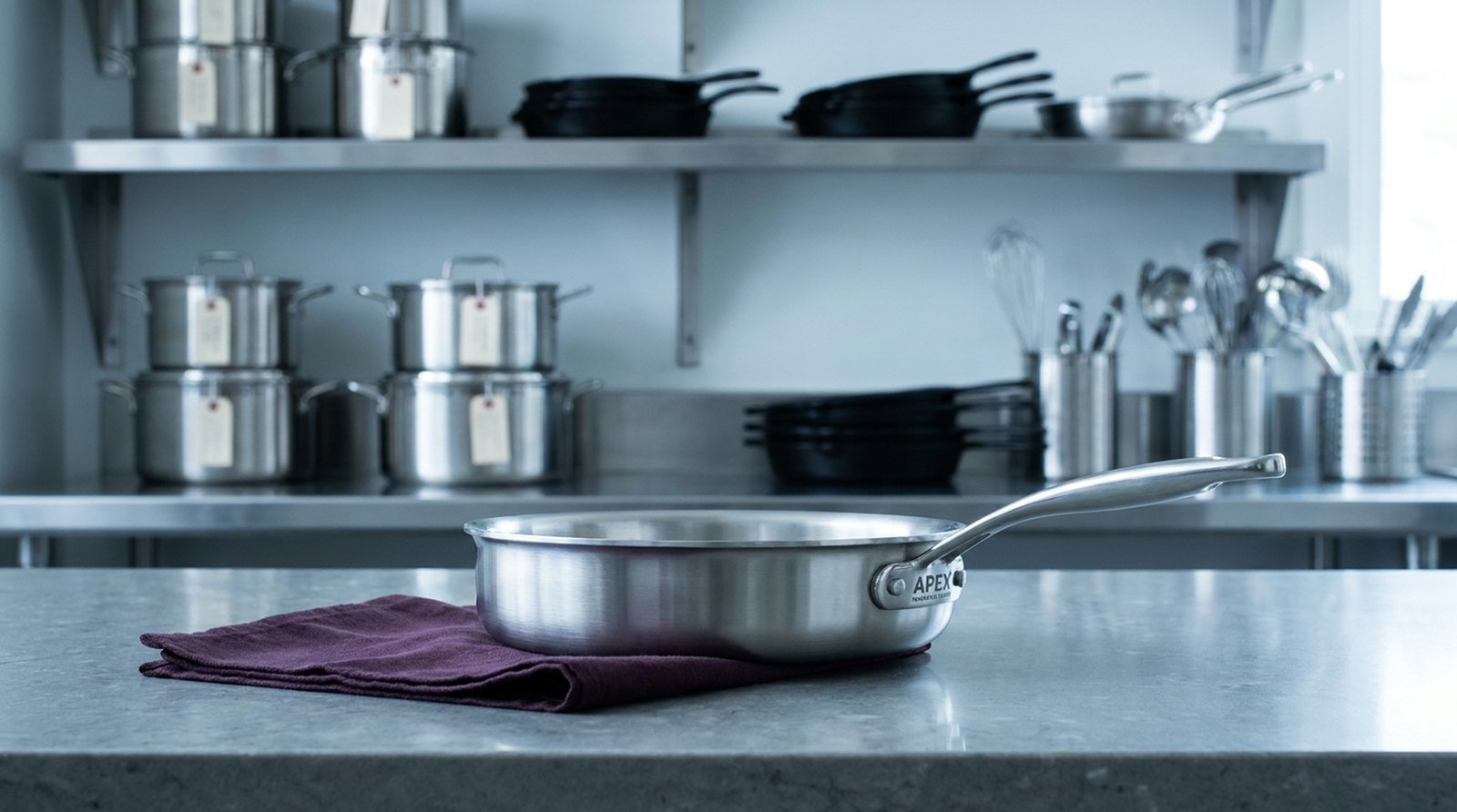A test-kitchen equipment room: brushed-steel shelving of tagged cookware above a honed-stone prep counter